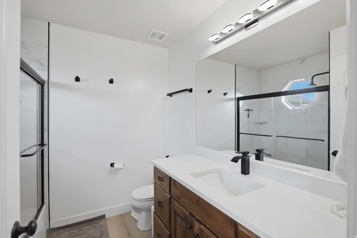 Spacious bathroom featuring a wood-finish vanity with an integrated sink, black hardware, a large mirror, a shower with a sliding glass door, and a shower window
