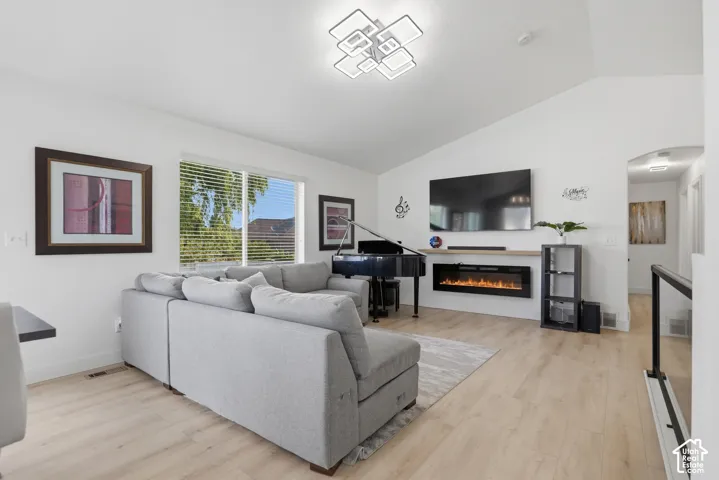 Vaulted ceiling, contemporary linear chandelier, light wood-finish flooring, electric fireplace with mantel, and a window with horizontal blinds