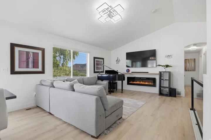 Vaulted ceiling, contemporary linear chandelier, light wood-finish flooring, electric fireplace with mantel, and a window with horizontal blinds