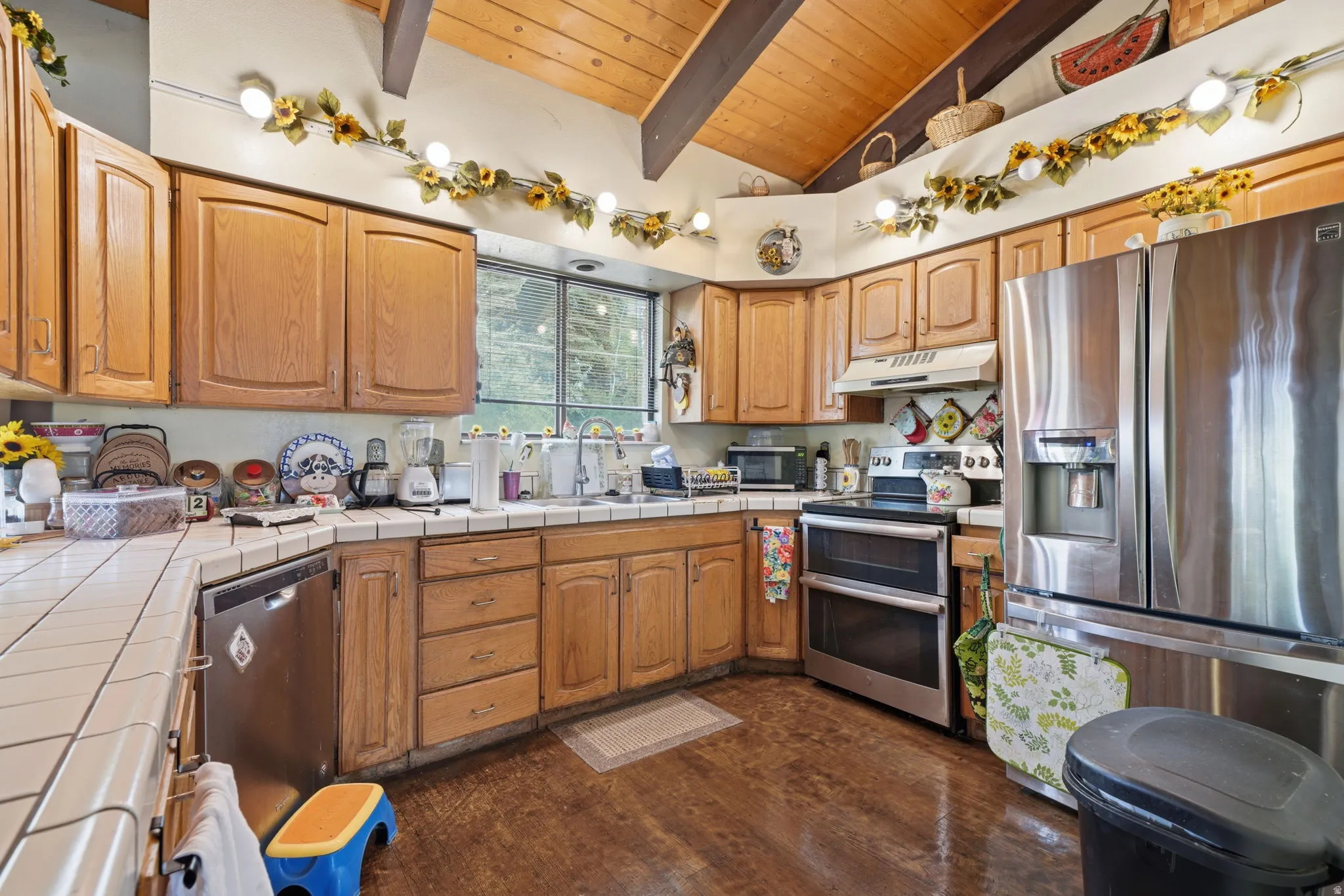 Kitchen featuring wood cabinetry, stainless steel appliances, tile countertops, wood-finish flooring, and a vaulted wood ceiling with exposed beams