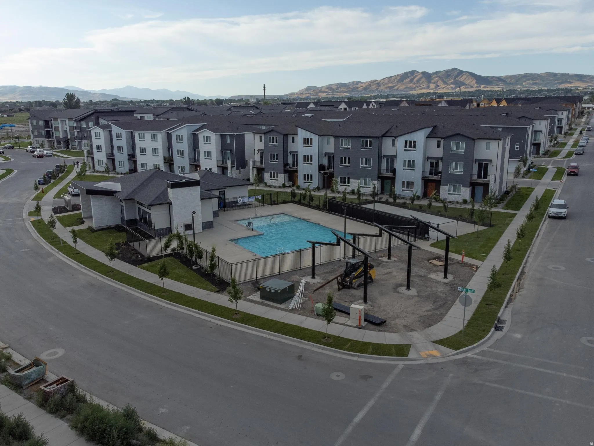 Modern residential buildings with dark roofs and light-colored siding, a swimming pool, and a surrounding paved area, all set against a distant mountain backdrop