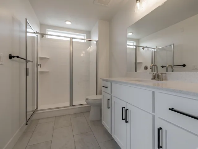 White shaker cabinetry with black hardware, double vanity with light-colored countertop, and a walk-in shower with glass enclosure