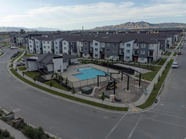 Modern residential buildings with dark roofs and light-colored siding, a swimming pool, and a surrounding paved area, all set against a distant mountain backdrop