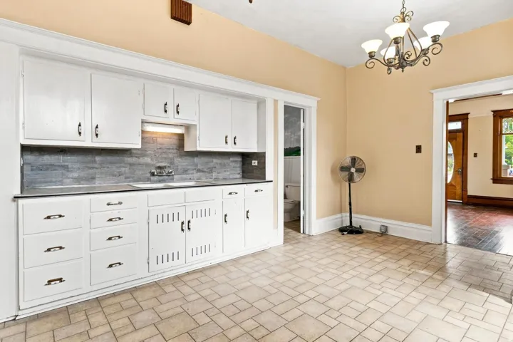 White cabinetry with dark hardware, gray tiled backsplash, dual basin sink, tiled flooring, and a decorative chandelier