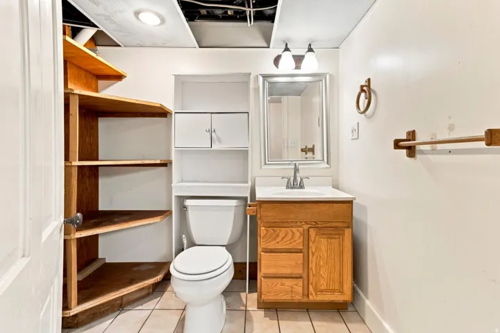 White walls with tile flooring, wood-finish vanity with a white countertop and chrome faucet, rectangular mirror with a dual-bulb vanity light, built-in shelving, and white toilet
