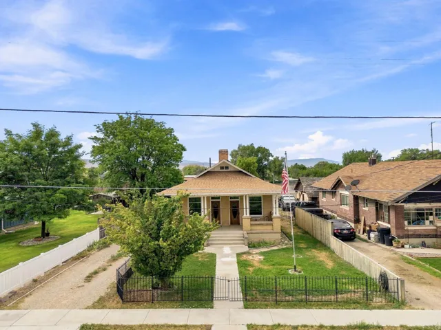 Gable roofline, covered front porch, front yard with mature trees, black wrought iron fence, and sidewalk access
