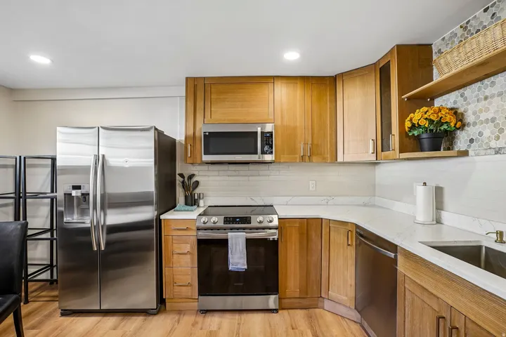 Wood-finish flooring, stainless steel appliances, light wood cabinetry, white stone countertops, and a mosaic tile accent wall
