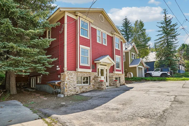 Red siding exterior with stone veneer foundation, multiple windows, covered entry, and mature evergreen landscaping