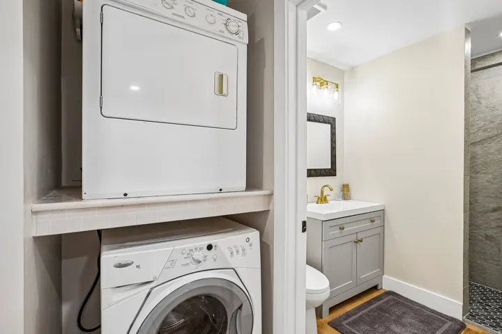Stackable laundry appliances, light gray vanity with white countertop, gold-finish faucet, framed mirror, and recessed lighting