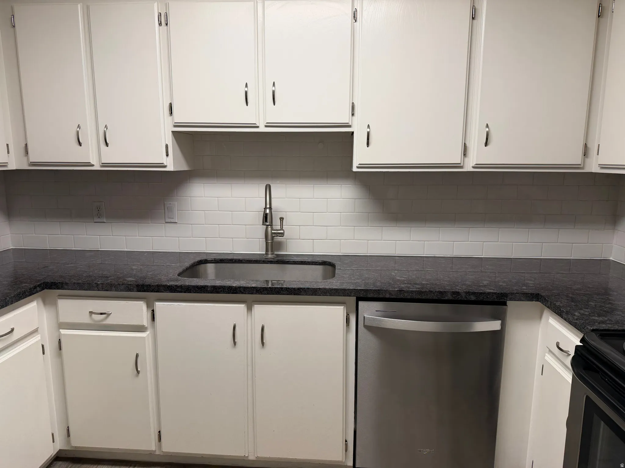 White cabinetry with brushed nickel hardware, dark speckled countertops, subway tile backsplash, and an undermount stainless steel sink with a high-arc faucet