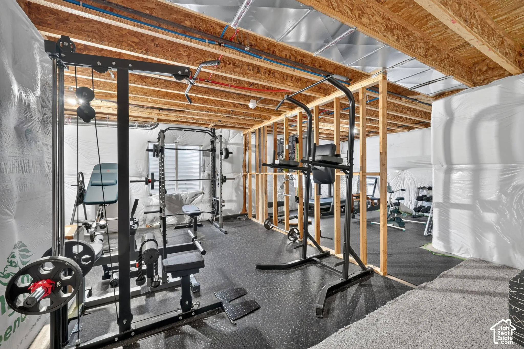 Unfinished basement with exposed ceiling joists and wood framing, featuring a rubber mat flooring section and a light-toned carpeted area
