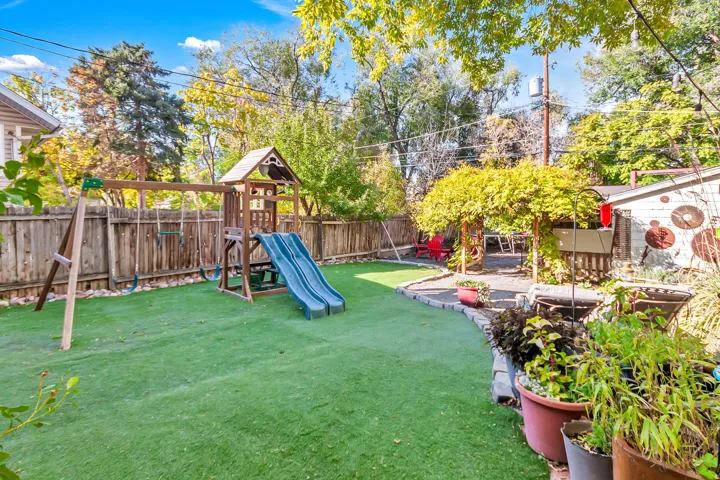 Expansive backyard featuring artificial turf, a wood privacy fence, a wooden play structure, a paved patio area, and mature trees