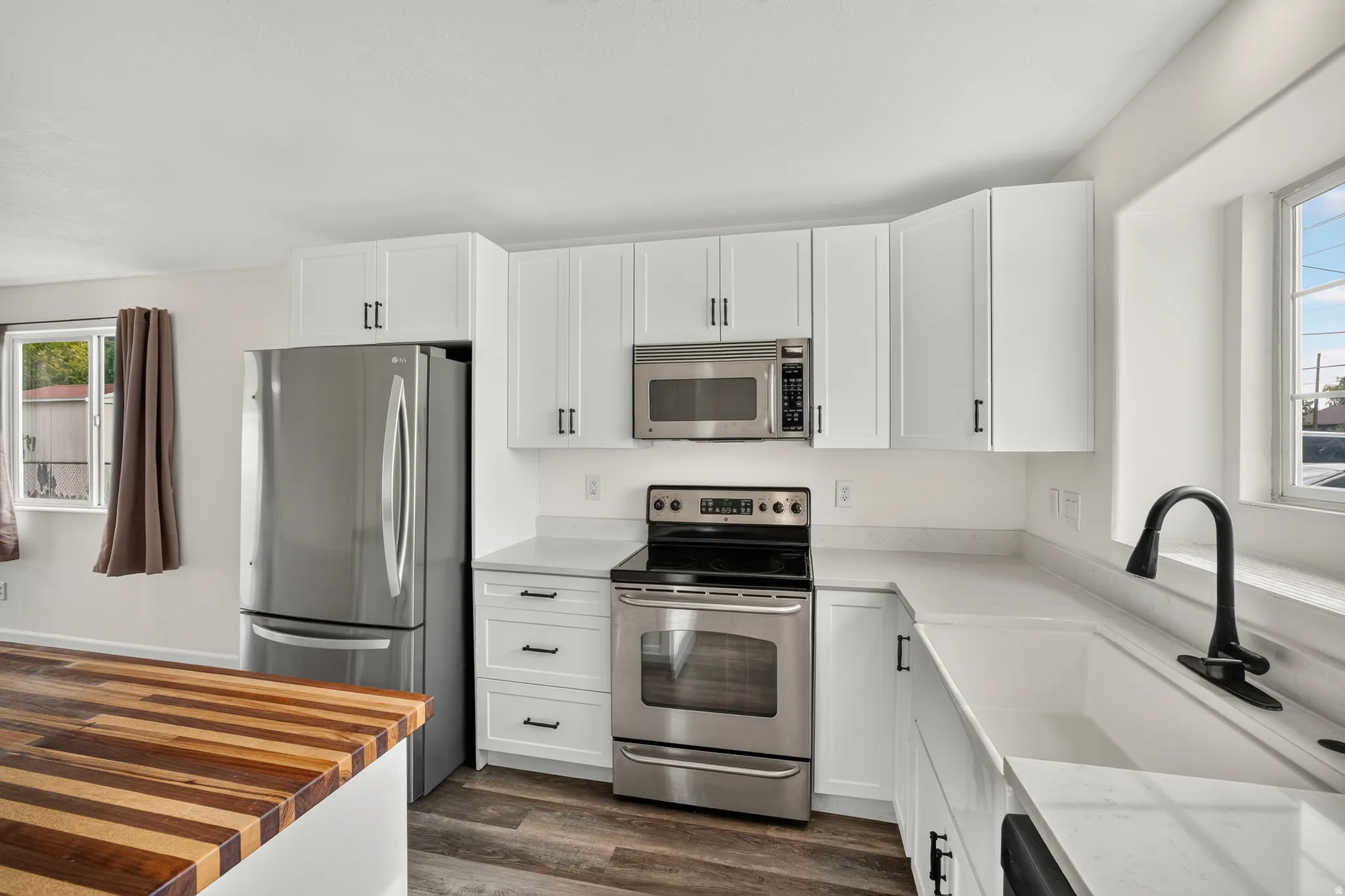 White shaker-style cabinetry with matte black hardware, stainless steel appliances, a white rectangular sink with a matte black faucet, and wood-finish flooring
