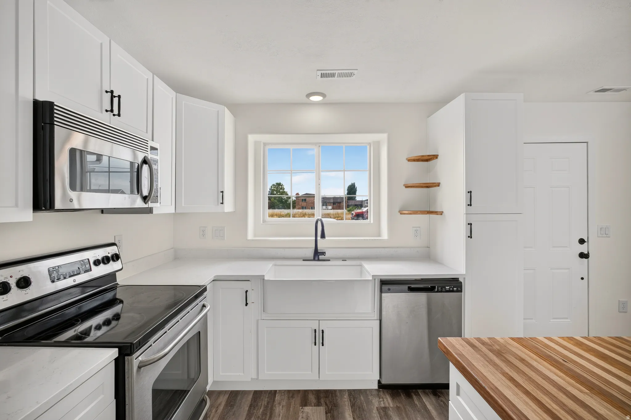 White shaker cabinetry, stainless steel appliances, farmhouse sink with matte black faucet, open wood shelving, and wood-finish flooring
