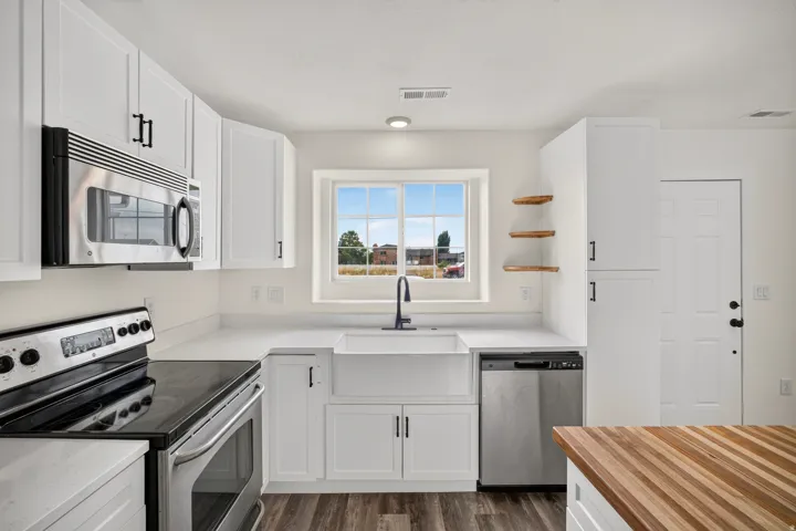 White shaker cabinetry, stainless steel appliances, farmhouse sink with matte black faucet, open wood shelving, and wood-finish flooring