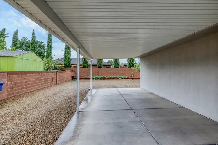 Covered patio with concrete flooring, supported by metal posts, a textured exterior wall, and a low brick retaining wall
