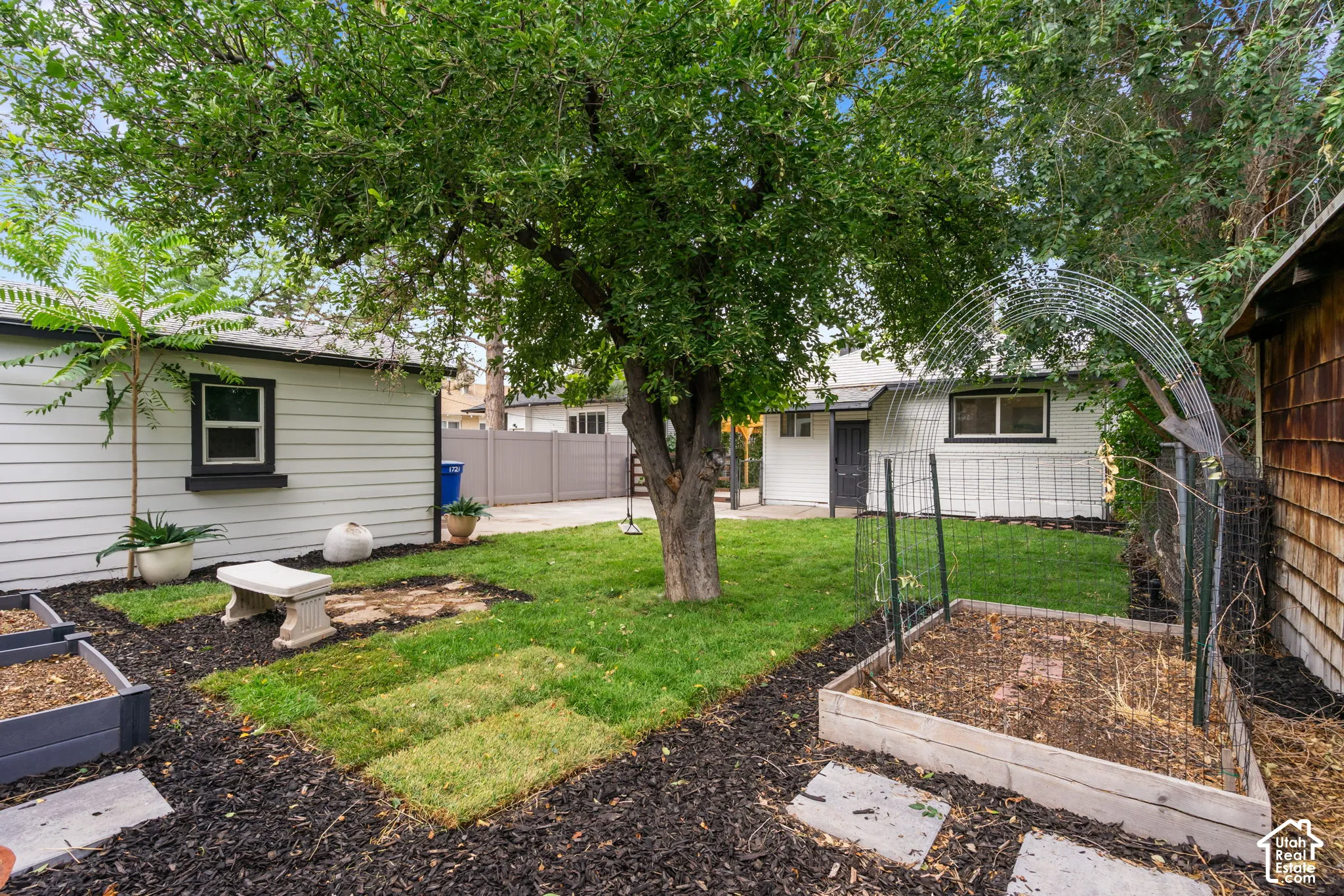 Expansive backyard featuring mature trees, raised garden beds, a stone bench, a paved walkway, and a privacy fence