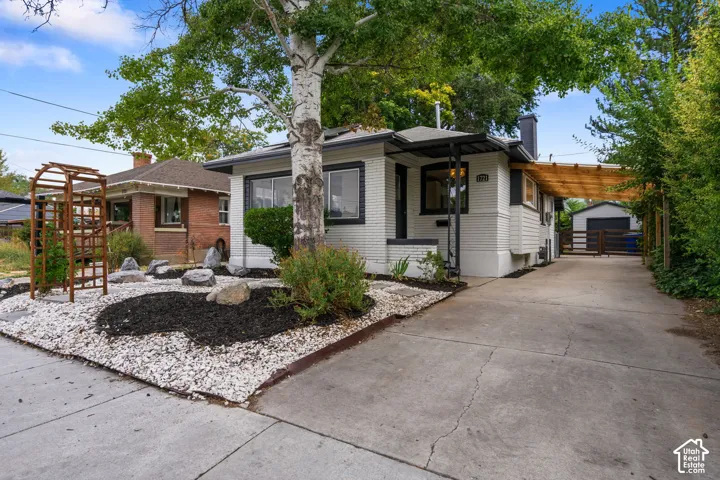White siding exterior with dark trim accents, a front porch with an awning, a wood pergola over the driveway, a detached garage, and a landscaped front yard with white gravel and dark mulch