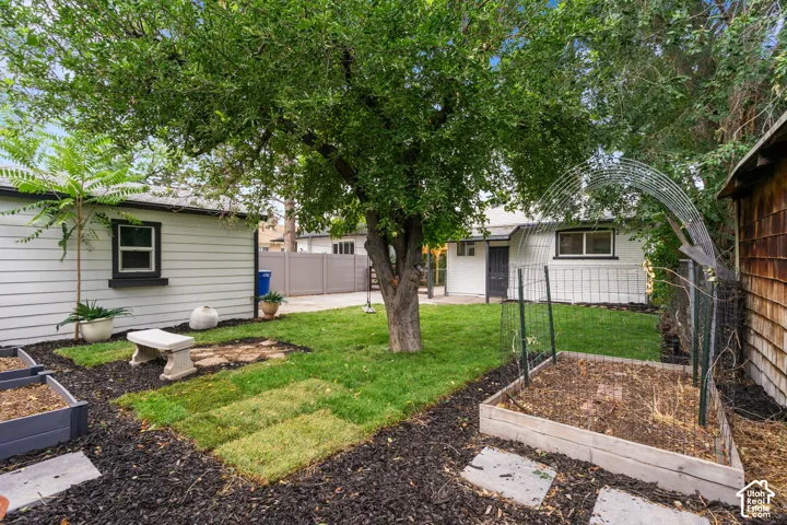 Expansive backyard featuring mature trees, raised garden beds, a stone bench, a paved walkway, and a privacy fence