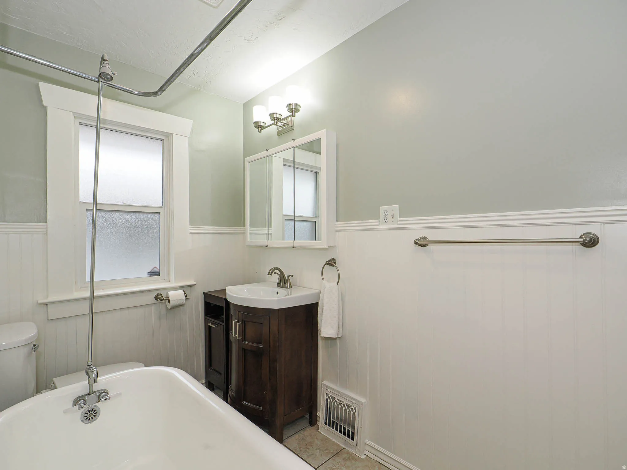 Spa-inspired bathroom featuring a freestanding soaking tub, dark wood vanity with integrated sink and brushed nickel fixtures, large framed mirror with sconce lighting, subway tile wainscoting, soft sage green upper walls, and natural light from a transom