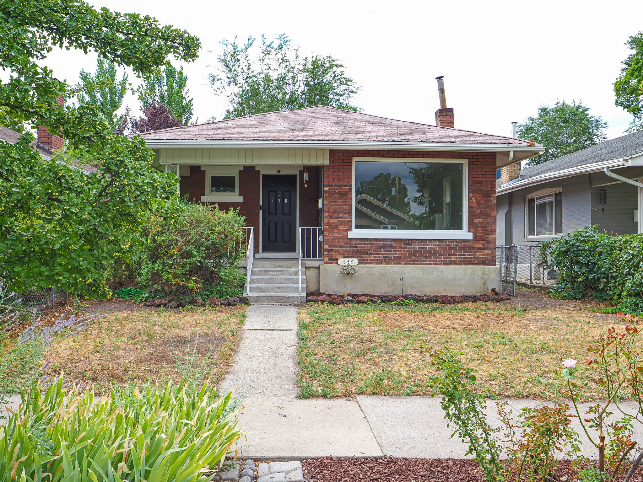 Charming brick ranch home featuring a large front-facing window, covered entry porch with concrete steps, and a manicured front lawn bordered by mature landscaping and foundation plantings