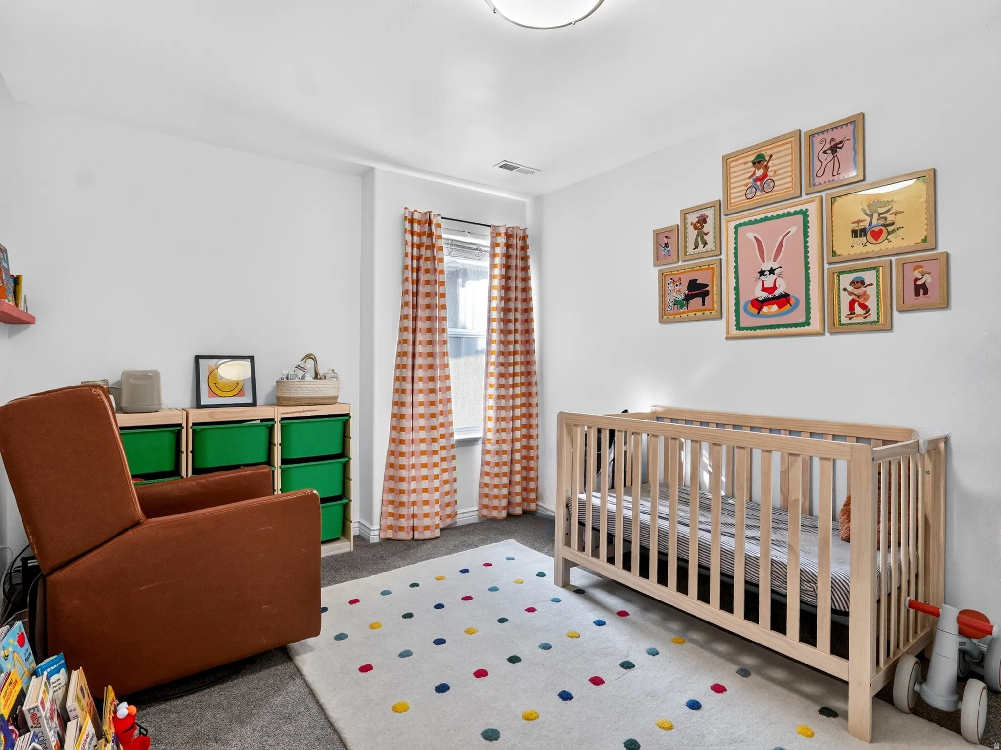 Bright nursery featuring soft white walls, natural light from multi-pane windows with patterned curtains, wooden crib with natural finish, green storage cubbies with integrated shelving, colorful area rug with geometric pattern, and a gallery wall of fram