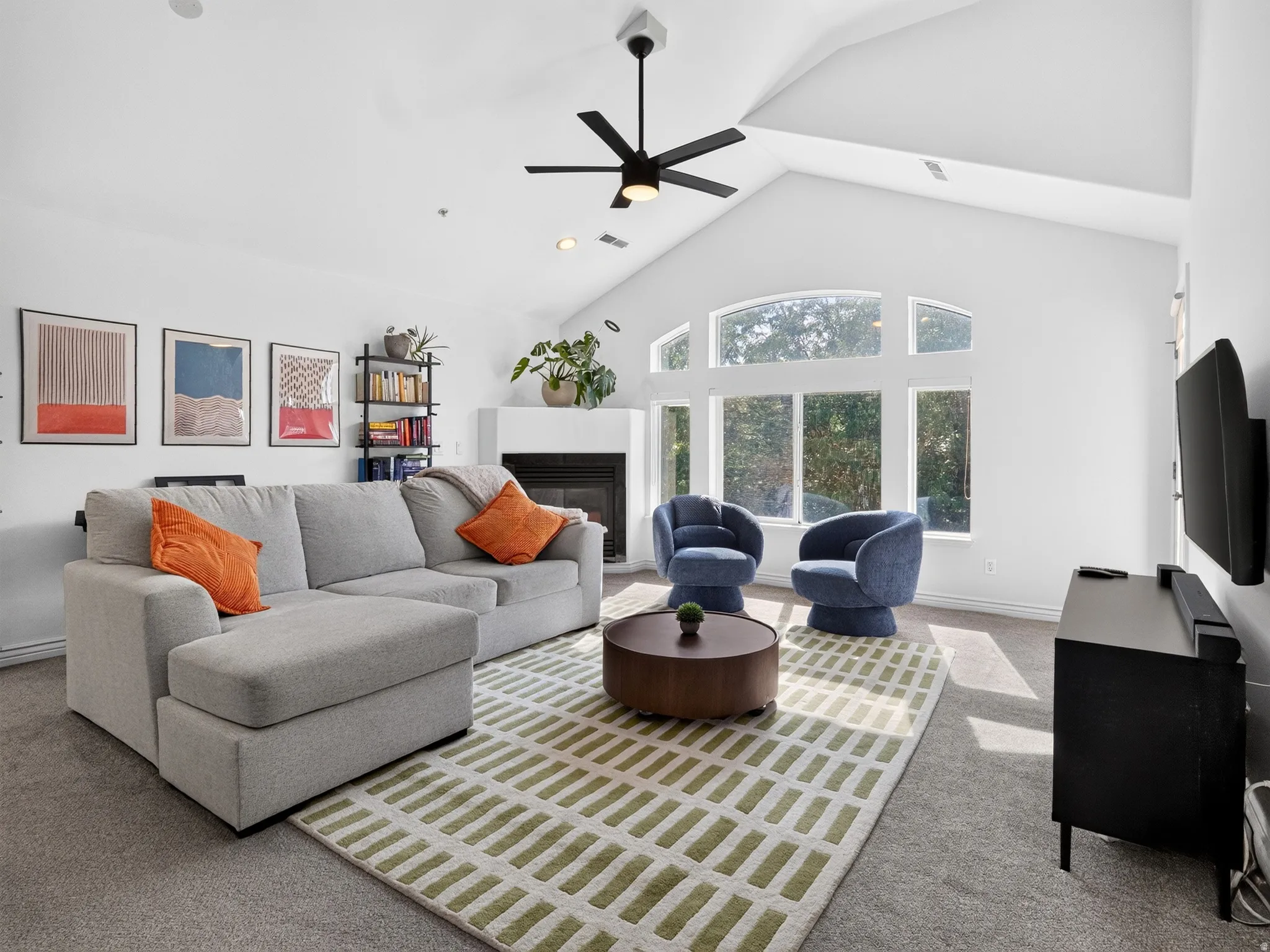 Bright, open living space featuring vaulted ceilings with ceiling fan, expansive multi-pane windows flooding the room with natural light, fireplace with dark surround, wide-plank wood-finish flooring, geometric patterned area rug, and contemporary dark ac