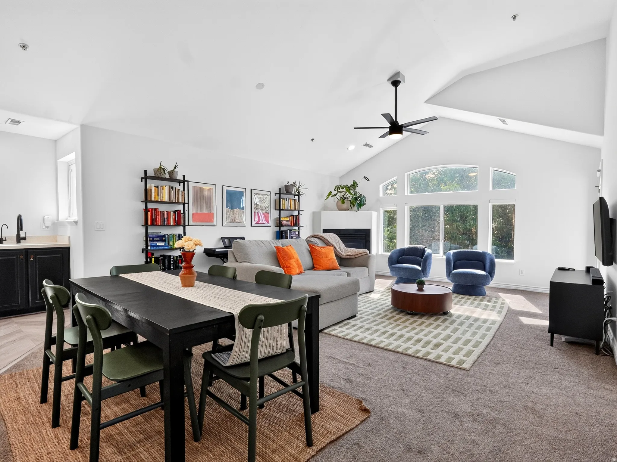 Open-concept living space anchored by a black dining table with seating for four, flowing into a generous great room featuring a gray upholstered sectional with navy accent chairs, ceiling fan, and abundant natural light from large arched windows