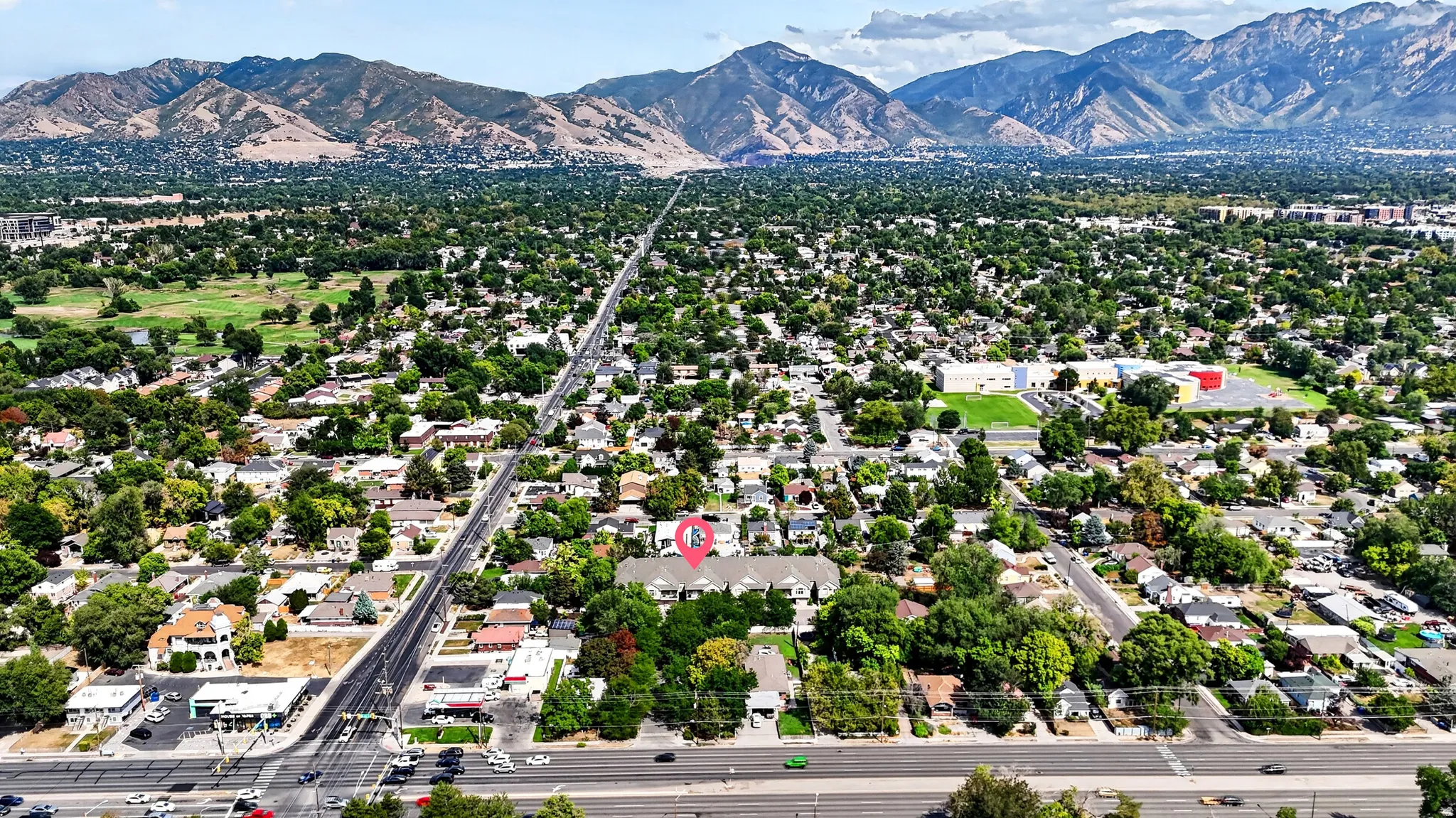 Aerial perspective showcasing a tree-lined residential community with mature landscaping throughout the neighborhood, a major thoroughfare providing direct access, and distant mountain vistas framing the property's setting