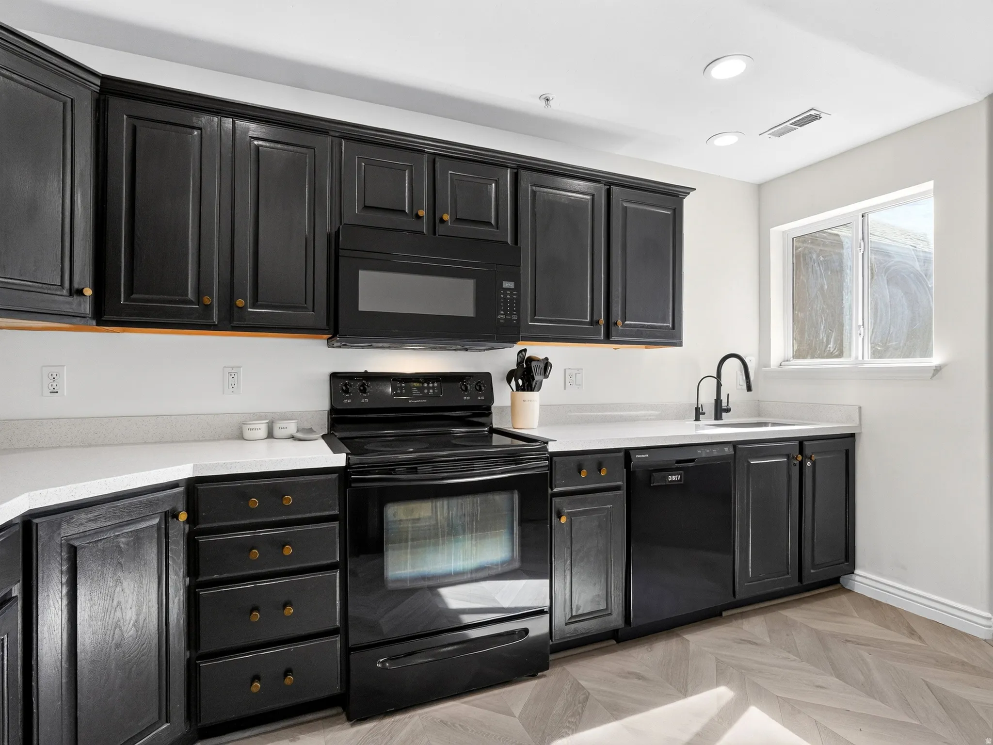 Contemporary kitchen featuring black cabinetry with brass hardware, stainless steel appliances including a slide-in range and built-in microwave, quartz countertops, an undermount sink with a gooseneck faucet, recessed ceiling lighting, and herringbone wo
