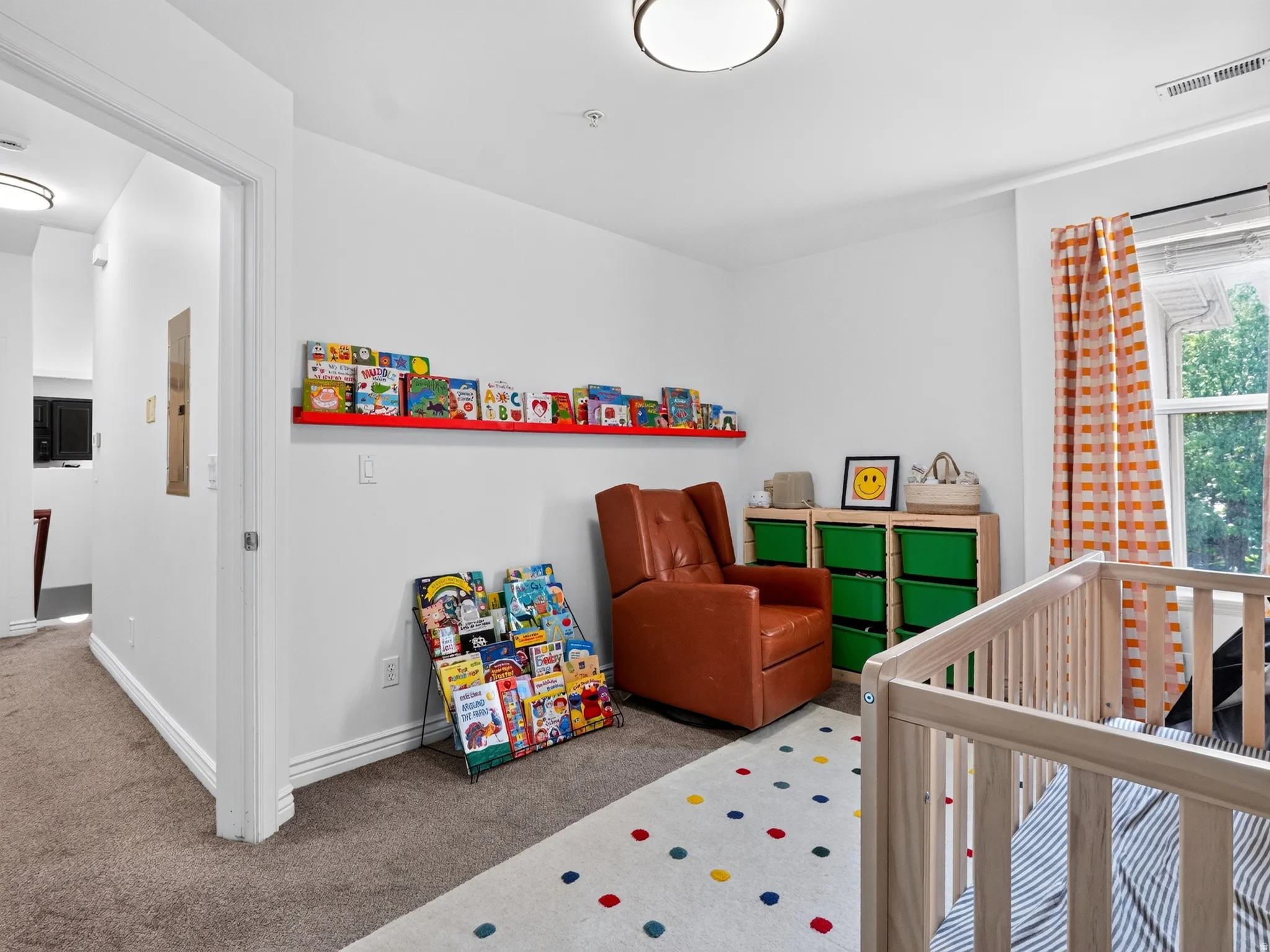 Bright nursery featuring soft carpeting with colorful polka-dot detailing, white crib with railings, built-in green storage cubbies, cognac-toned upholstered chair, red wall-mounted shelving for books and décor, large window with orange patterned curtain