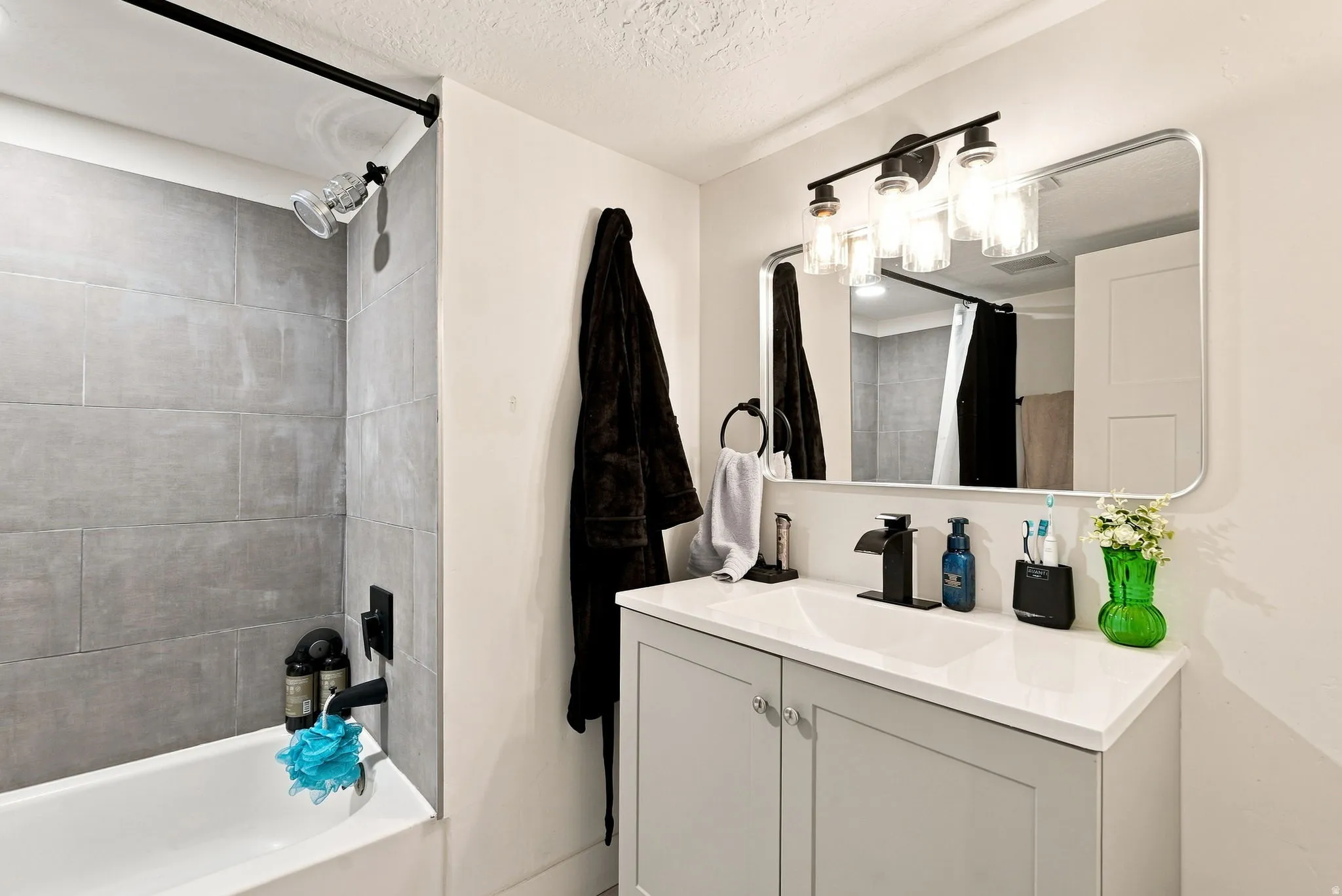 Bathtub with gray tile surround, matte black shower fixtures, vanity with light gray cabinetry, white countertop, and a rectangular mirror with integrated lighting