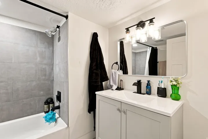 Bathtub with gray tile surround, matte black shower fixtures, vanity with light gray cabinetry, white countertop, and a rectangular mirror with integrated lighting