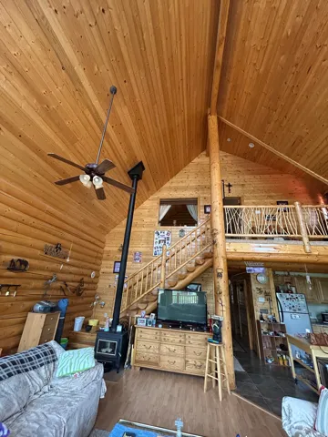 Vaulted wood-paneled ceiling with exposed wood beams, interior log wall construction, wood-finish flooring, wood staircase with log railing, and a black wood-burning stove