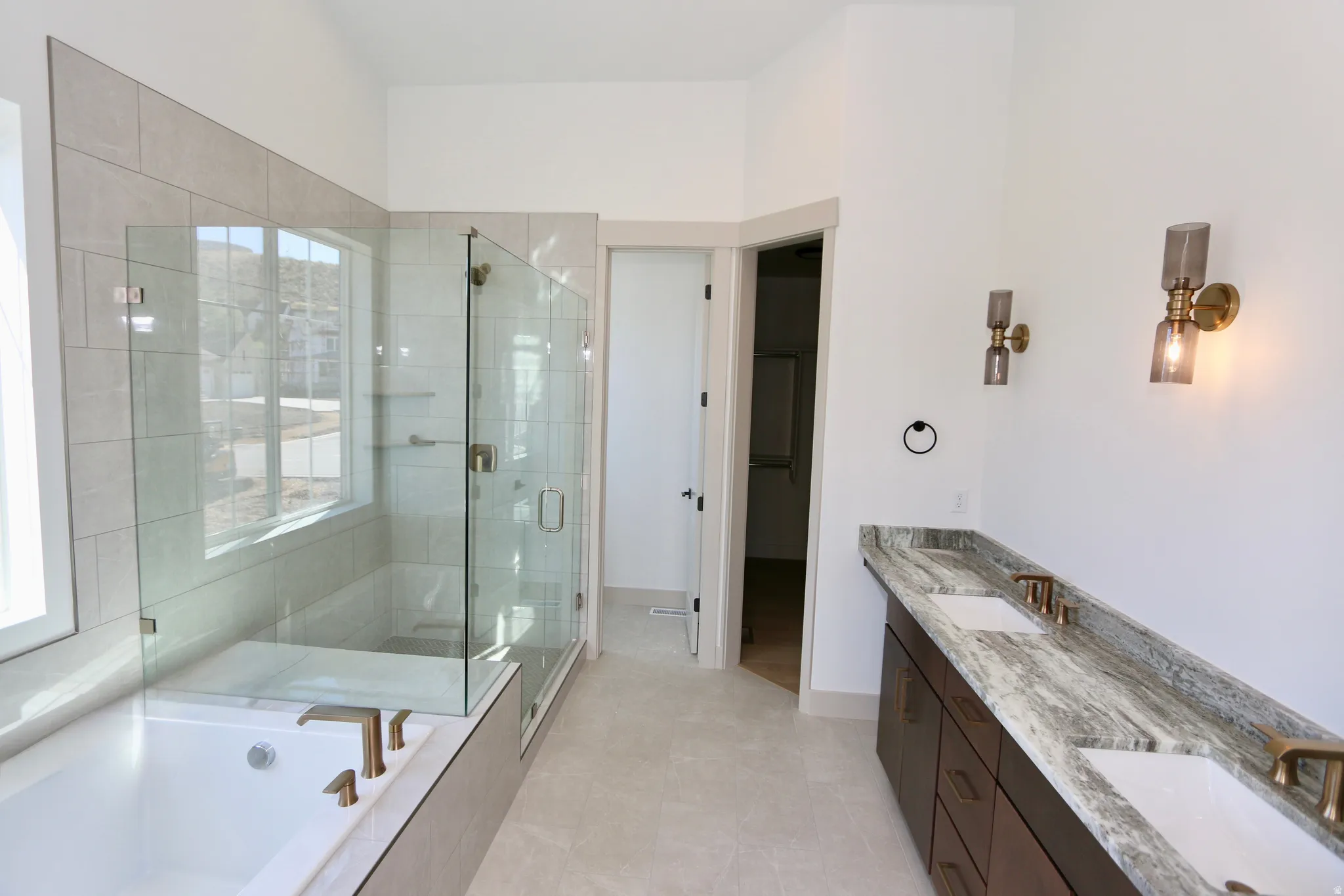 Large master-bath with dual vanity, quartzite countertop, soaking tub, euro-glass-enclosed shower with bench,, wall-mounted light fixtures, and tile flooring
