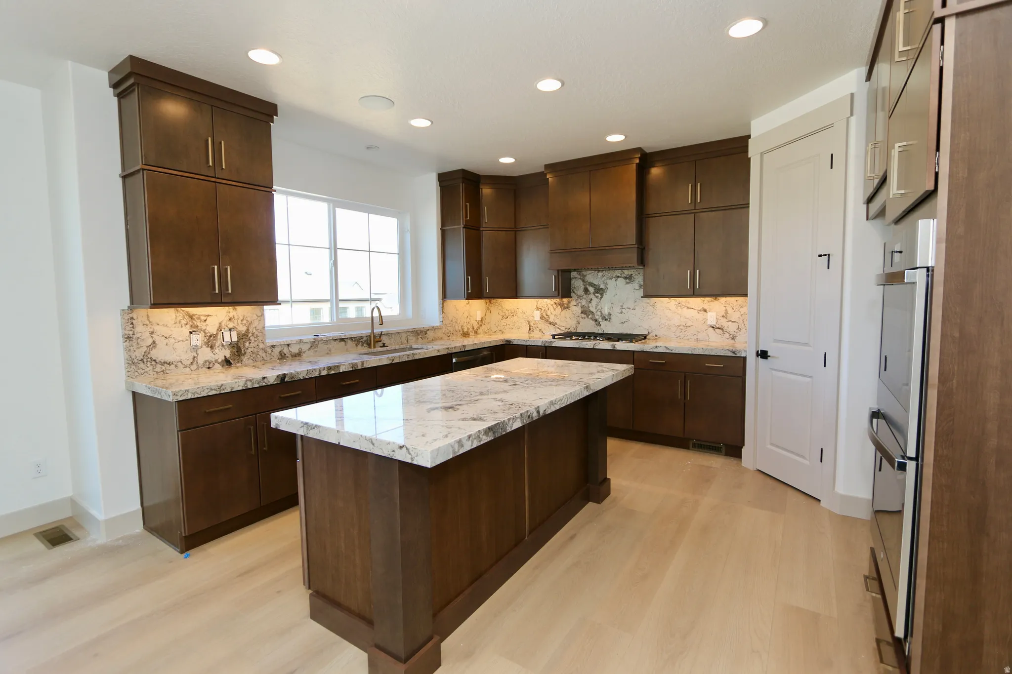 Kitchen featuring a center island, recessed lighting, wood-finish cabinetry, light-toned flooring, stone countertops with a matching backsplash and a large sink window