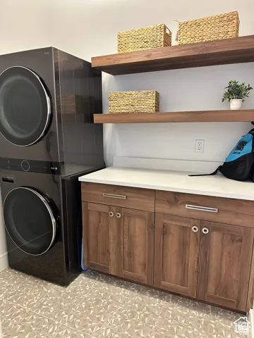 Laundry area featuring stacked appliances, wood-finish floating shelves, and wood-finish cabinetry with a white countertop