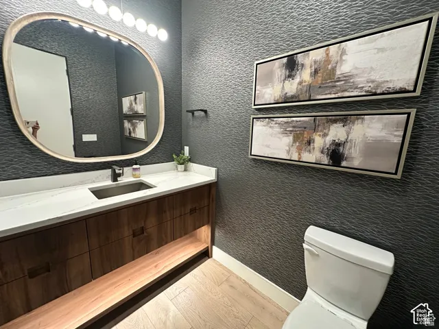Textured dark wall covering, wood-finish flooring, floating wood vanity with integrated lighting, white countertop, and contemporary oval mirror with integrated lighting