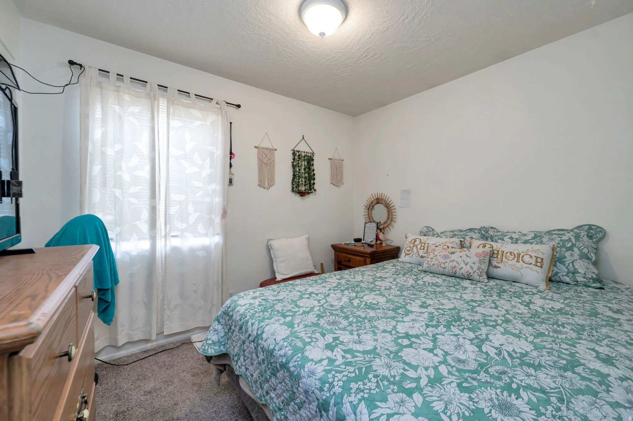 Bright secondary bedroom featuring a wood-finish dresser, carpet flooring, and sheer window treatments allowing natural light