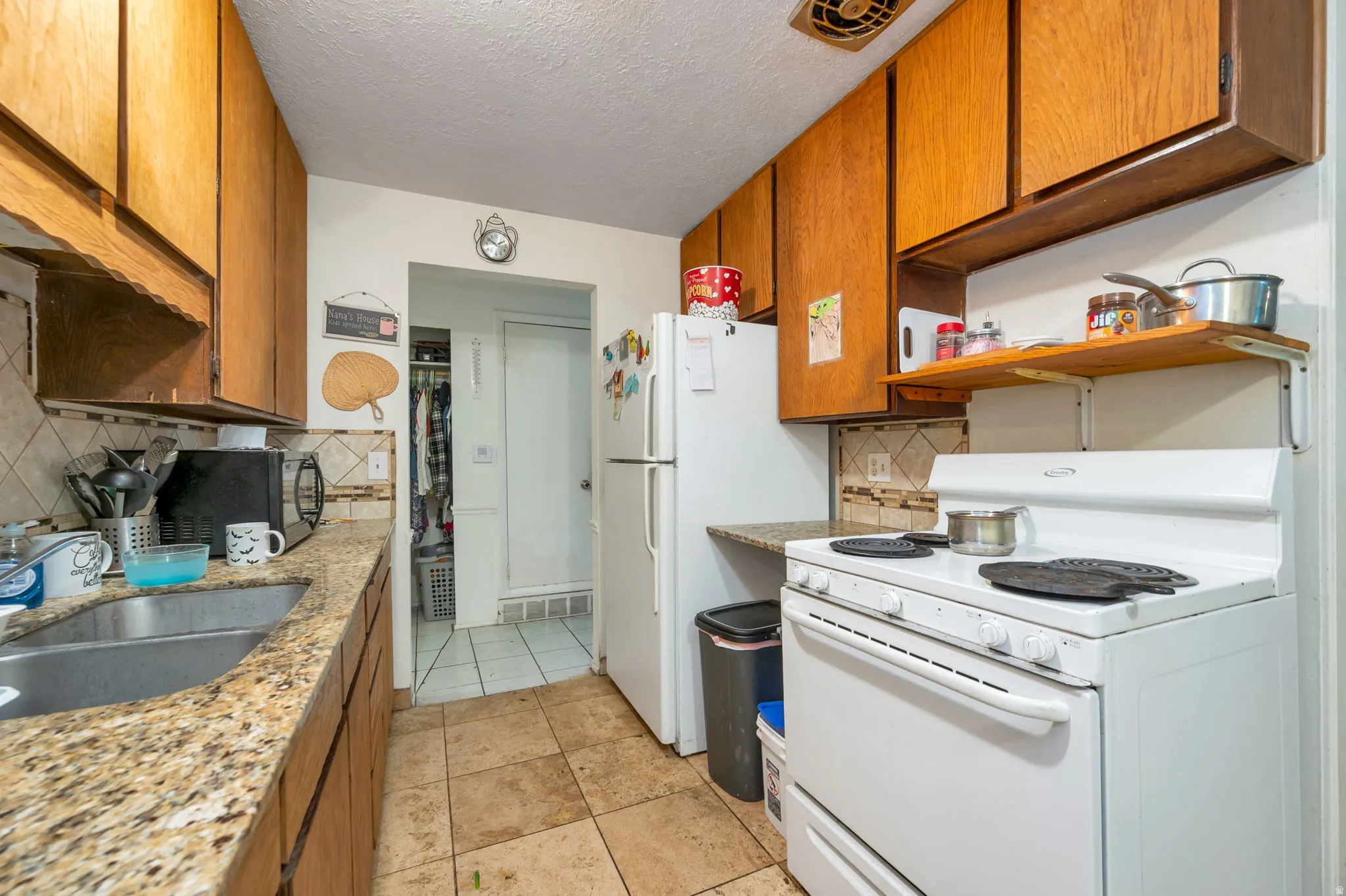 Galley kitchen featuring warm wood cabinetry with open shelving, speckled countertops, white electric range, stainless steel sink, and ceramic tile flooring throughout