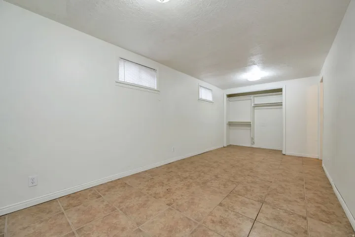 Neutral-toned floor tiles, white walls, two windows with blinds, and an open closet with shelving and hanging rods