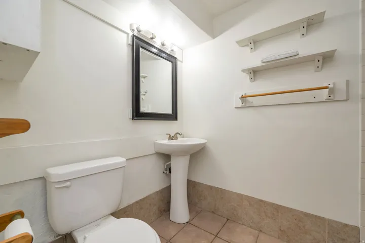 Compact bathroom featuring a pedestal sink, white porcelain toilet, tile flooring, and framed mirror with integrated lighting