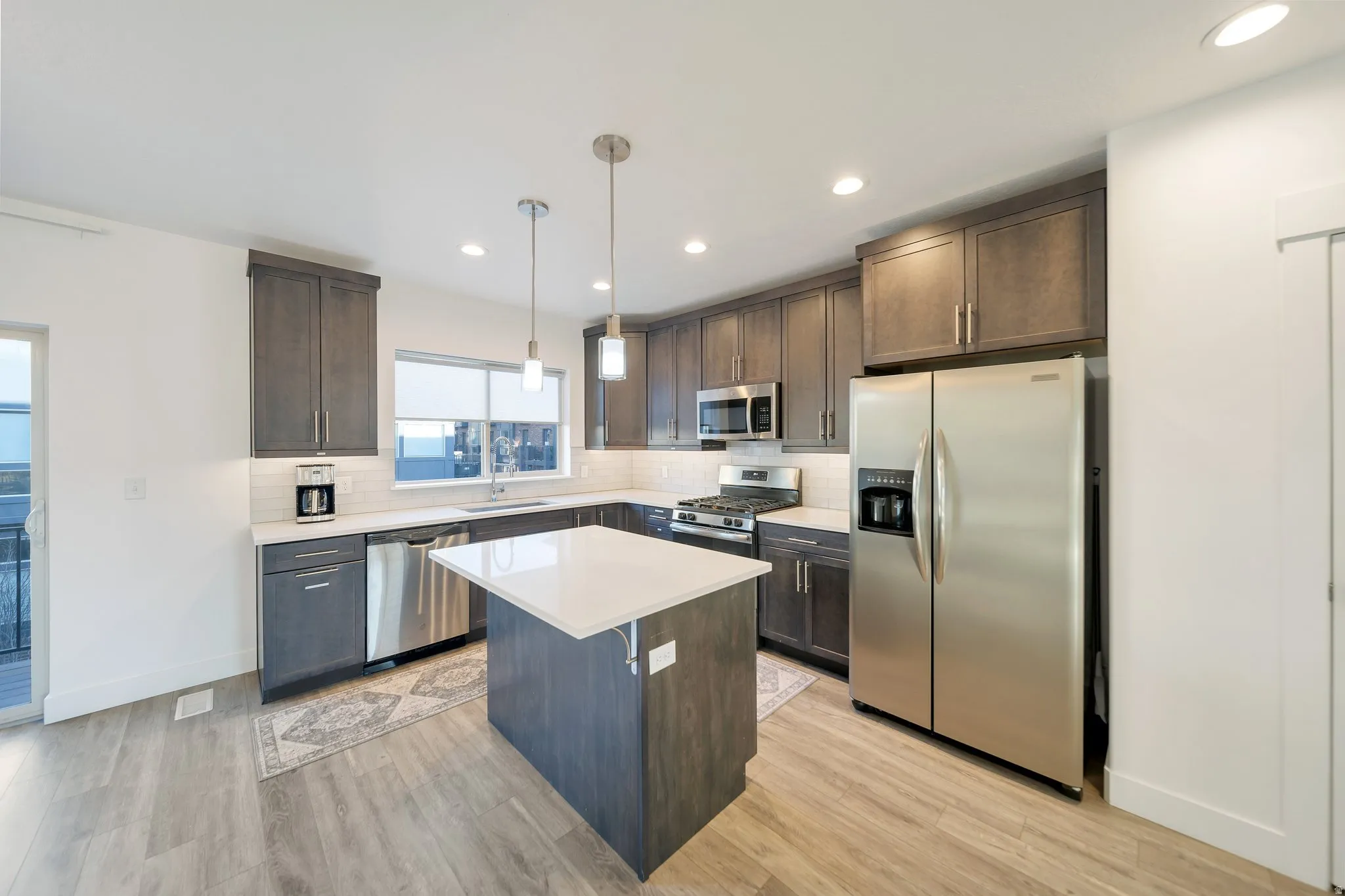 Kitchen featuring wood-finish flooring, dark wood cabinetry, stainless steel appliances, a white subway tile backsplash, and a central island with a white countertop