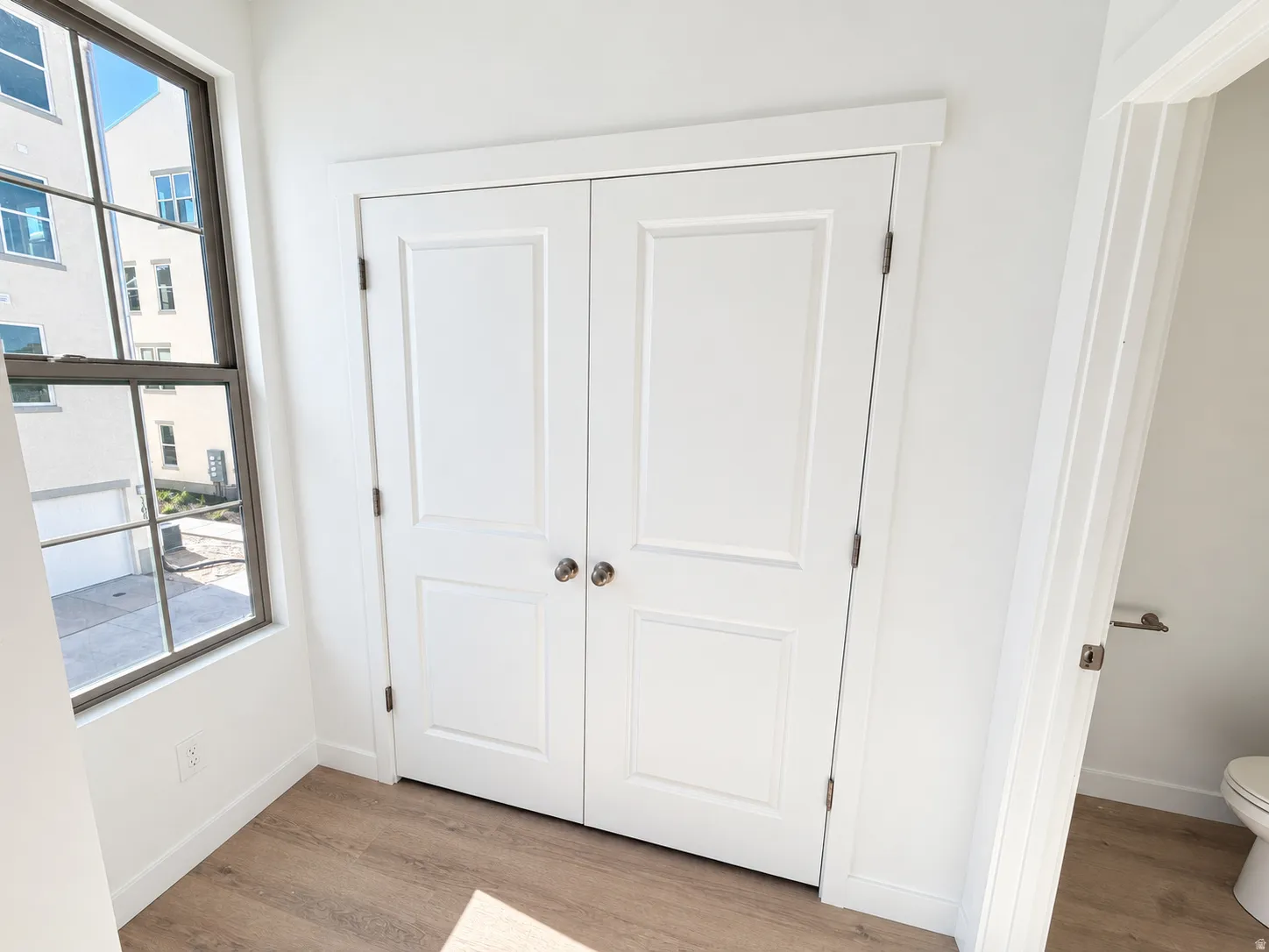 Tall, double-pane window with dark trim, modern paneled doors, light wood-finish flooring, and crisp white walls