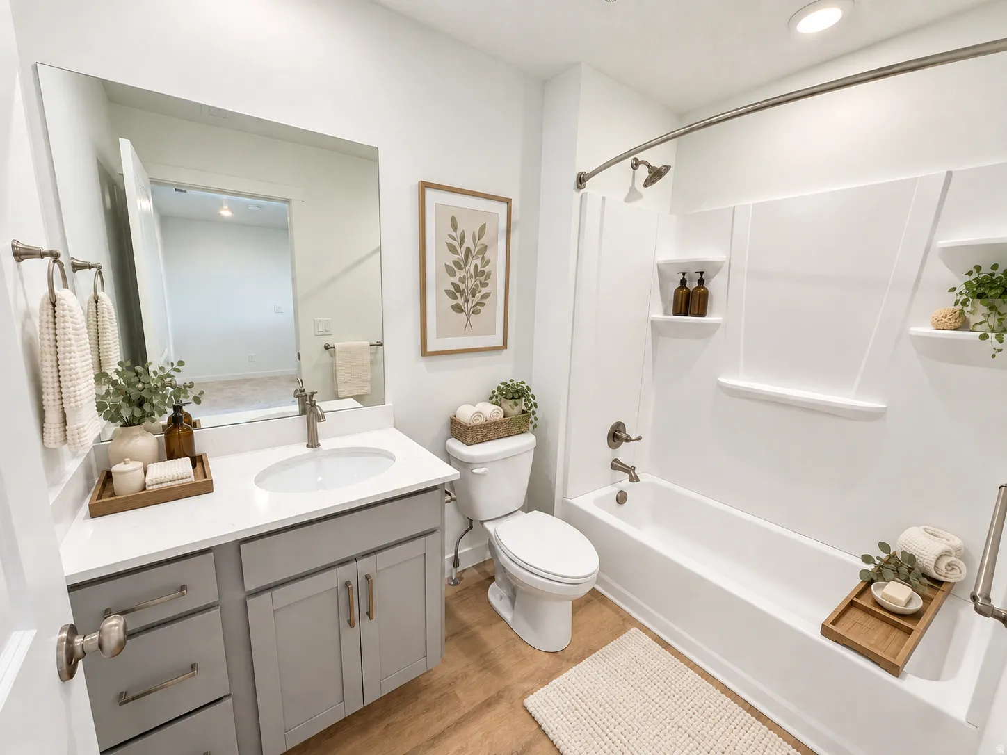 White vanity with a single basin sink and brushed nickel faucet, large frameless mirror, wood-finish flooring, white bathtub with integrated shelving, and a toilet