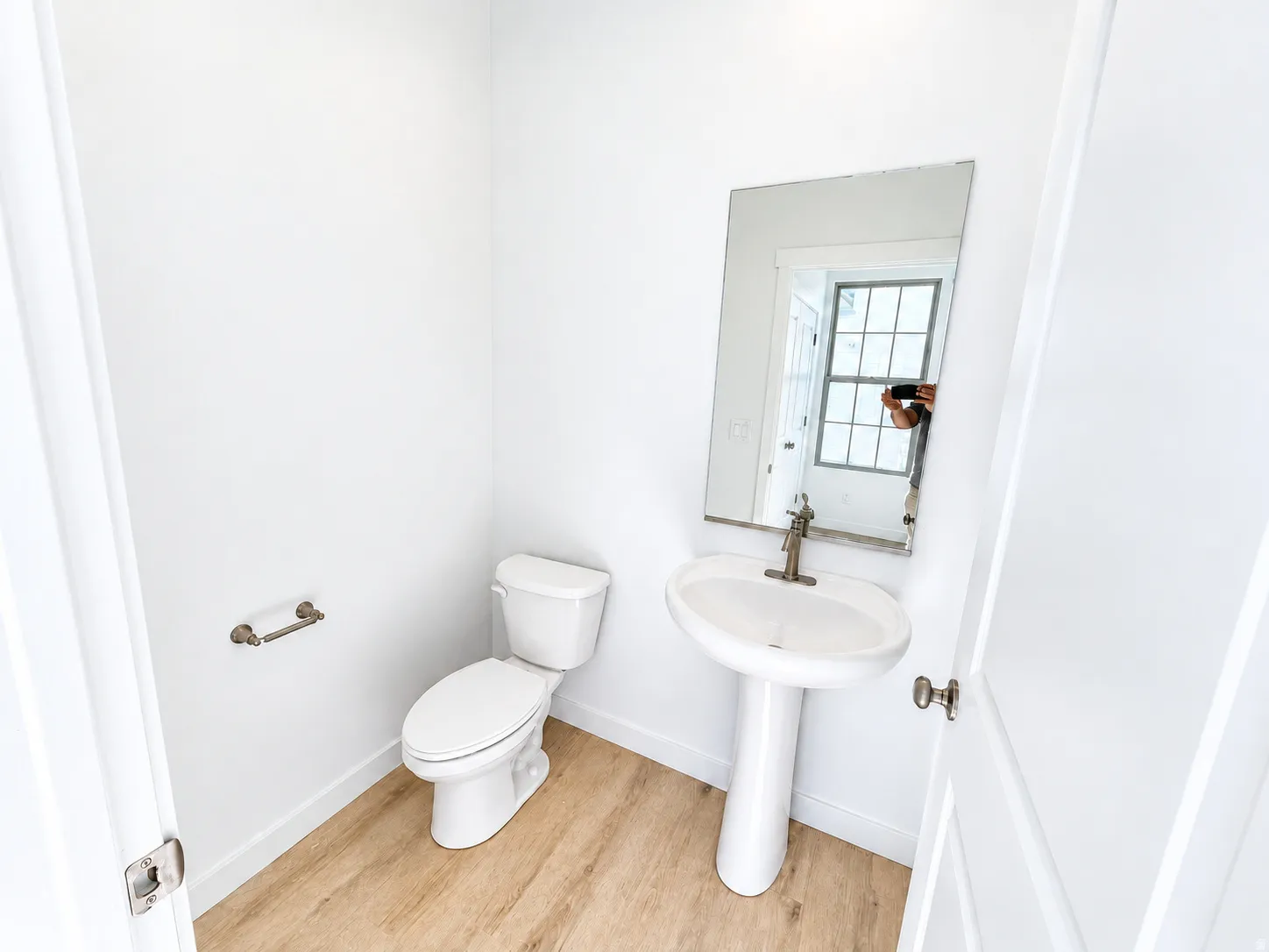 Pedestal sink with a single-handle faucet, rectangular wall mirror, toilet, and wood-finish flooring