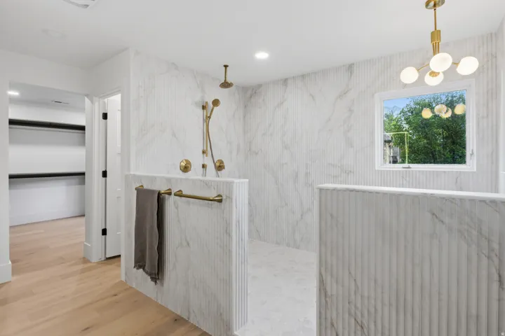 Walk-in shower with full-height fluted and veined tile walls, gold-tone fixtures including a rainfall showerhead, and a window for natural light