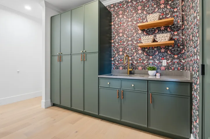 Light wood-finish flooring, built-in green cabinetry, integrated sink with brass faucet, two floating wood shelves, and patterned wall covering