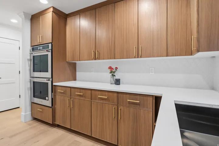 Wood-finish cabinetry with golden hardware, white countertops, stainless steel double wall ovens, and a white diamond-patterned backsplash