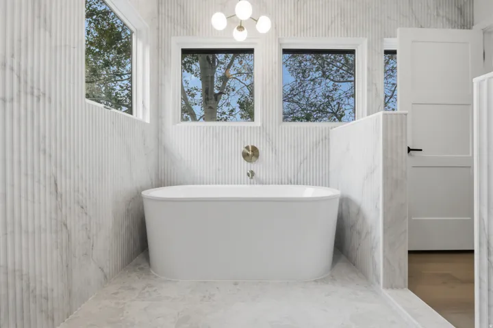 Freestanding soaking tub with a wall-mounted brass fixture, surrounded by fluted wall tile and two windows